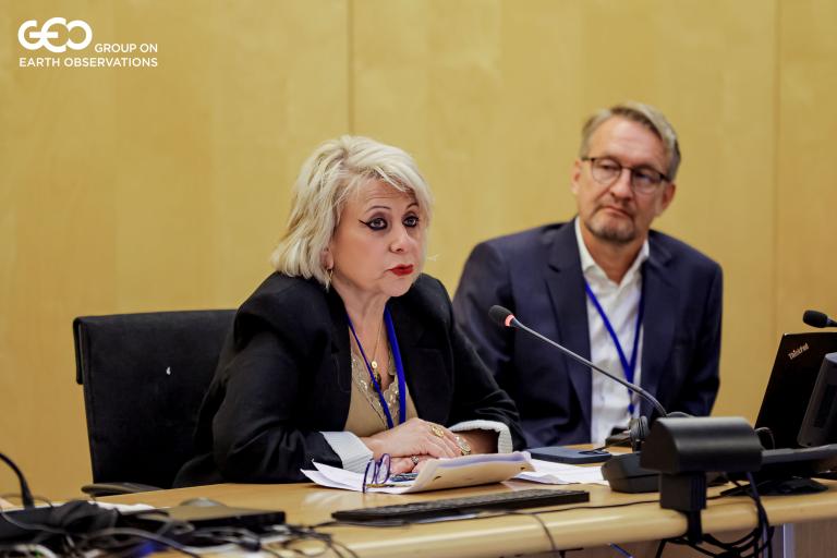A woman speaks into a microphone at a conference table, while a man sits beside her. Both have conference badges. GEO Group on Earth Observations logo is in the corner.
