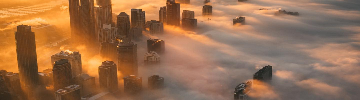 Aerial view of a city skyline at sunrise with tall buildings emerging through a thick layer of fog and golden sunlight illuminating the scene.