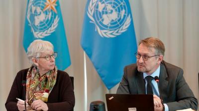 Two people sit at a table with microphones, engaged in discussion. Two United Nations flags are visible in the background.