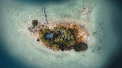 A small, oval-shaped island with trees, a single building, and a wooden dock, surrounded by shallow, clear water, viewed from above.