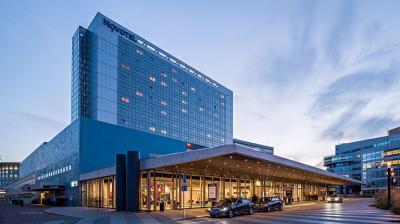 Large modern hotel building with a "Novotel" sign, glass windows, and a covered entrance where cars are parked; city buildings are visible in the background at dusk.