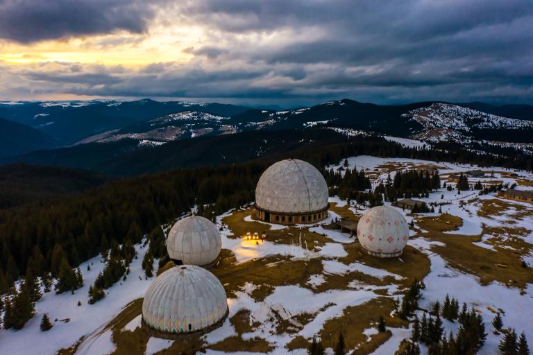 Aerial view of several large geodesic dome structures on a snowy, mountainous landscape under a cloudy sky at sunset.