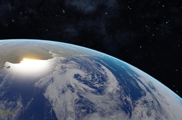 View of Earth from space showing clouds over the ocean, with sunlight illuminating part of the land near the planet's curved horizon against a dark space background.