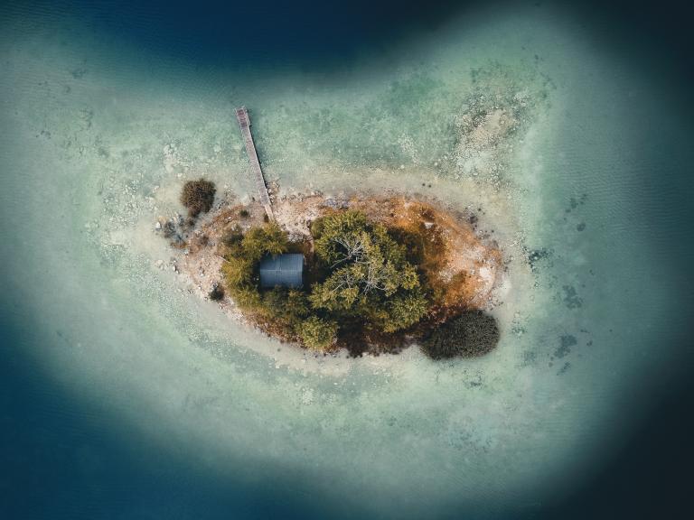 A small, oval-shaped island with trees, a single building, and a wooden dock, surrounded by shallow, clear water, viewed from above.