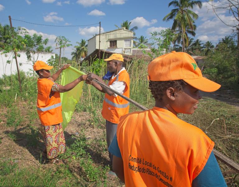 Three people in orange vests and hats work outdoors, holding a green bag and a tool, with a house and palm trees in the background.