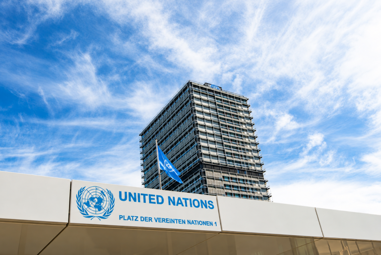 United Nations building with logo and flag under a blue sky with clouds; address “Platz der Vereinten Nationen 1” visible on the sign in front.